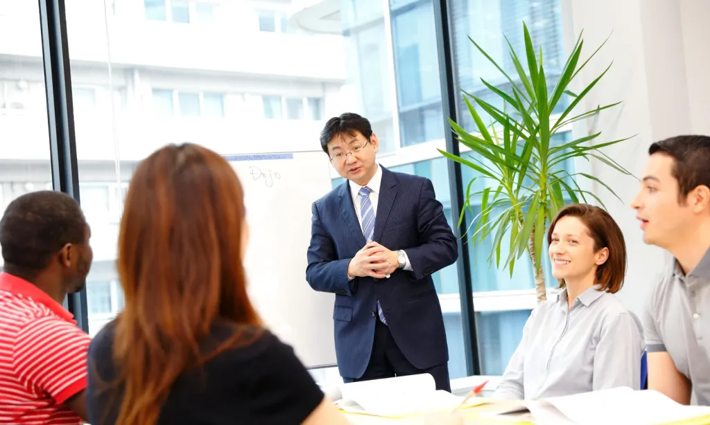 A professor in a suit teaches a diverse group of business students at a table. The whiteboard behind him says "Dojo".