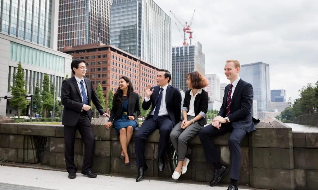 Five diverse business professionals chat casually while sitting on a stone wall in front of a modern city skyline.