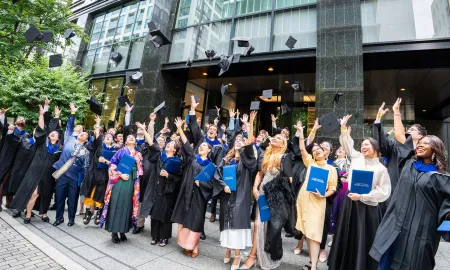 GLOBIS business school graduates joyfully throw their caps into the air in celebration outside a modern building.