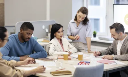 Diverse business students collaborate at a table with notes and coffee cups as a standing woman guides the discussion.