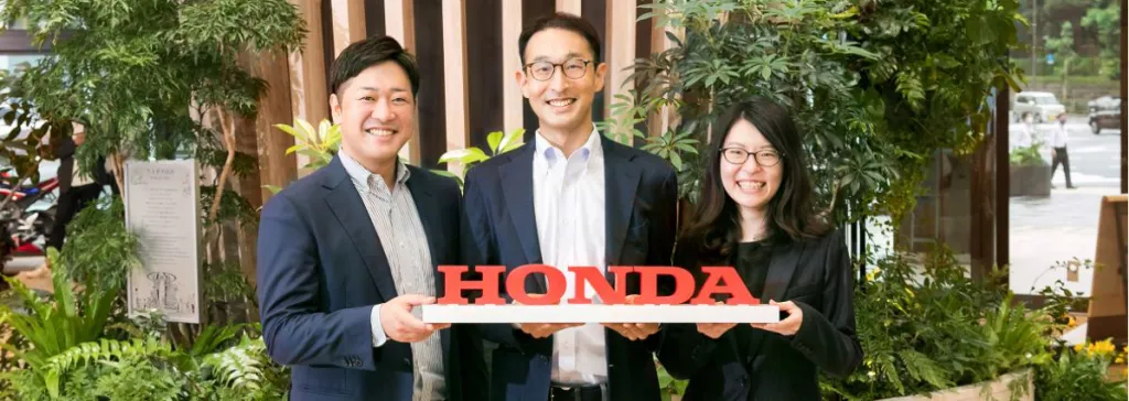 Three smiling professionals holding a red 3D Honda logo sign in a bright, plant-filled atrium.
